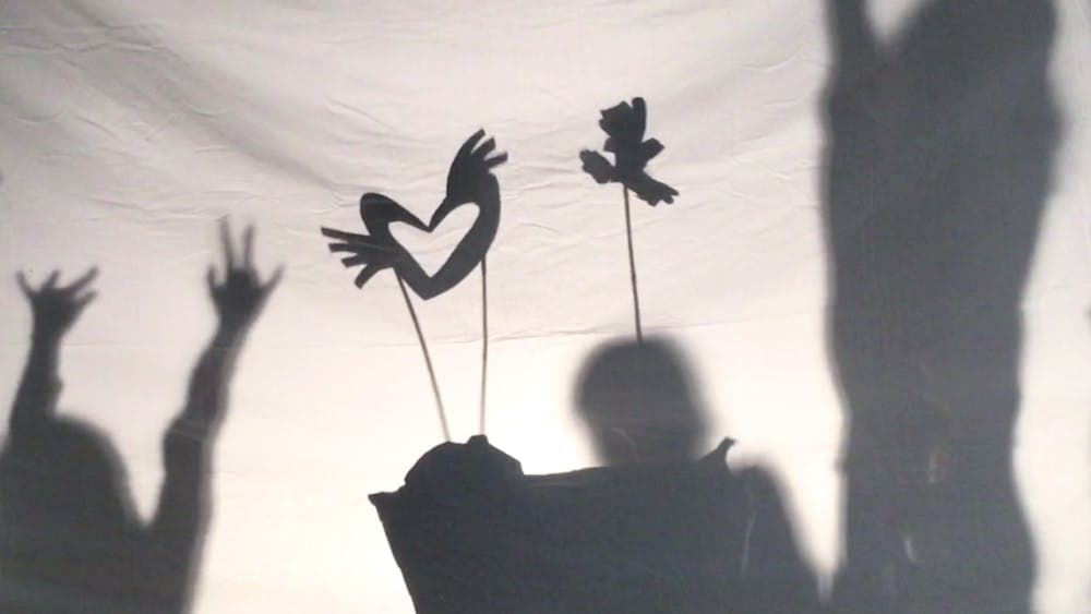 Shadow puppets move across a screen as the outlines of children dancing are visible on either side