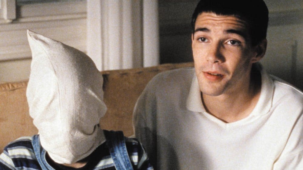 Man wearing a white polo next to a figure with a white pillowcase on their head
