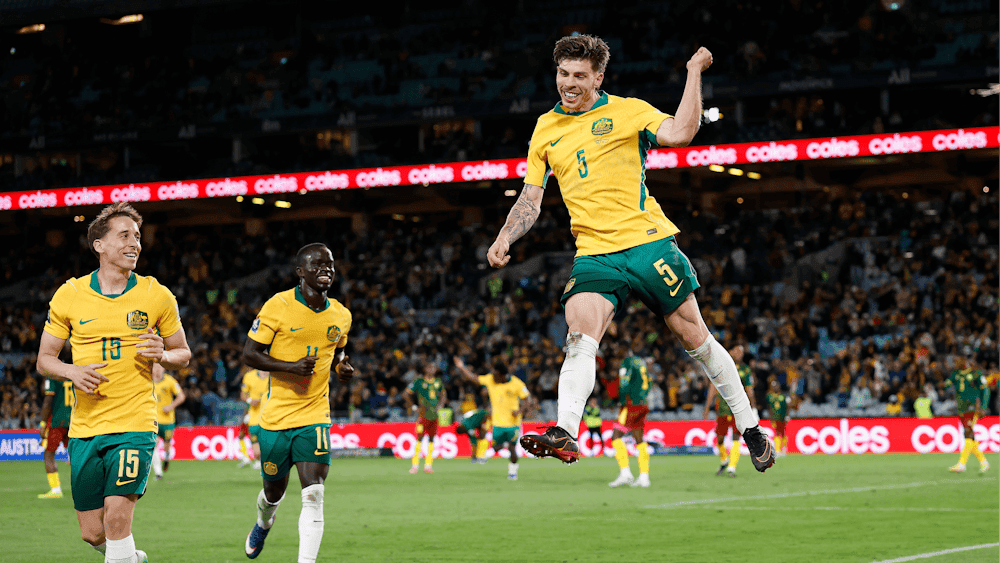 Soccer players in yellow kits celebrate a goal on a stadium pitch as teammates run in behind