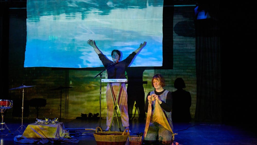 Two performers on stage with textiles, projected water imagery, and musical instruments.