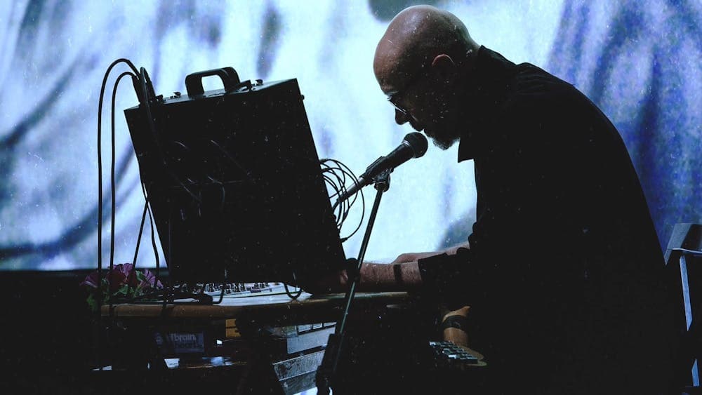 Performer sits at audio equipment, speaking into a microphone before a glowing blue projection.